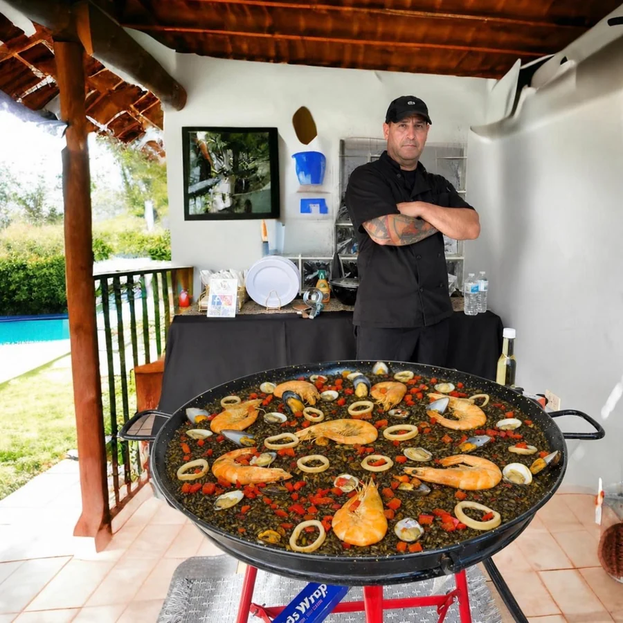 El chef Ángel de brazos cruzados detrás de una paella negra de mariscos recién hecha en un evento